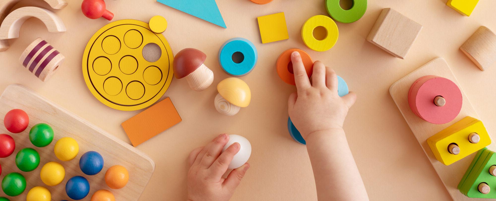 A child's hands are playing with colorful wooden educational toys, including geometric shapes, stacking blocks, and puzzle pieces on a beige surface.